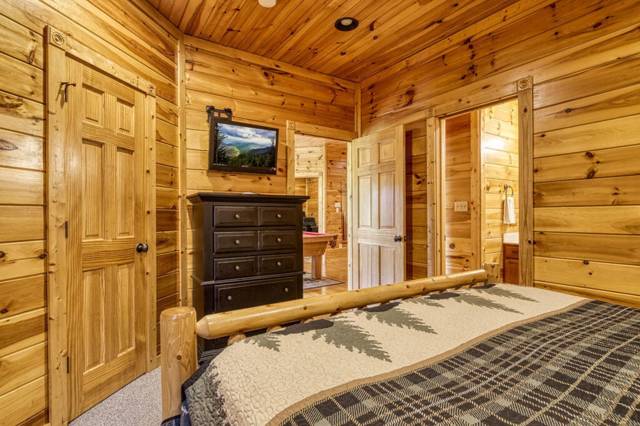 A wooden cabin interior with a bed, dresser, and a television mounted on the wall.