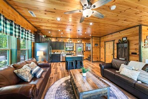 Un acogedor interior de cabaña con techo de madera, una zona de estar central con una mesa de centro, una cocina con taburetes y un gran ventanal.