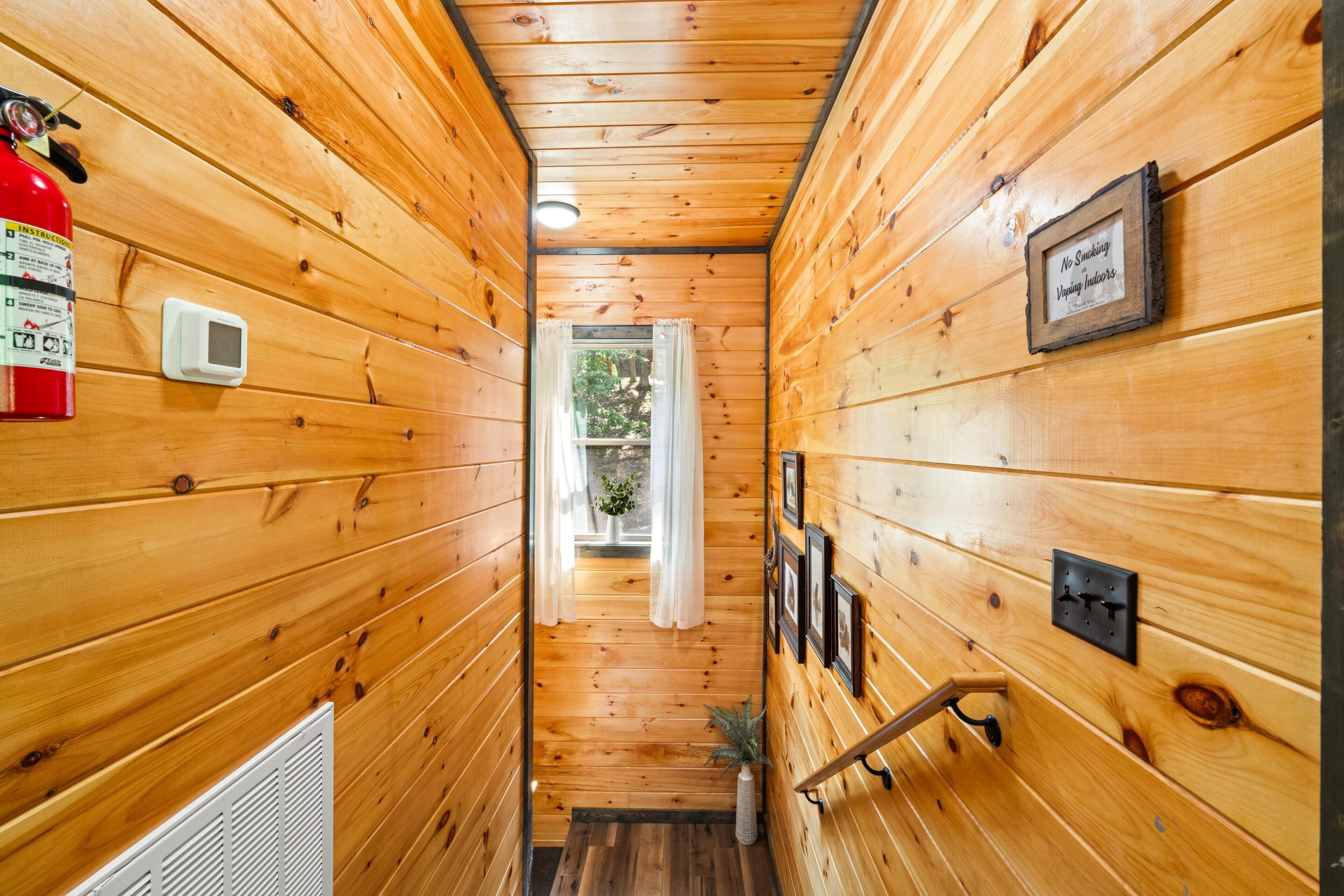 A wooden interior hallway with a fire extinguisher, a smoke detector, a small window with curtains, and a framed sign.