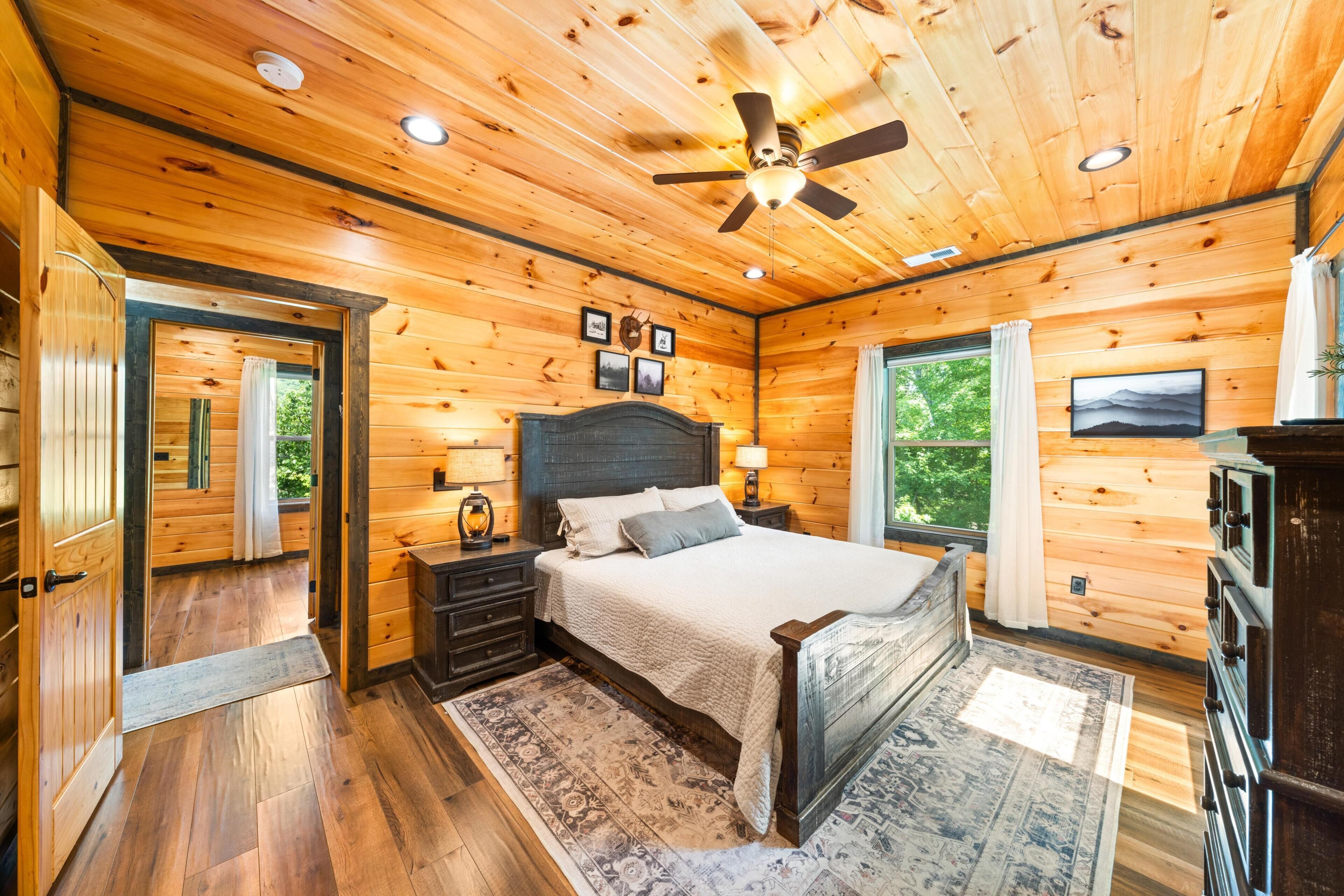 A wooden cabin bedroom with a bed, nightstands, a ceiling fan, and a large window.
