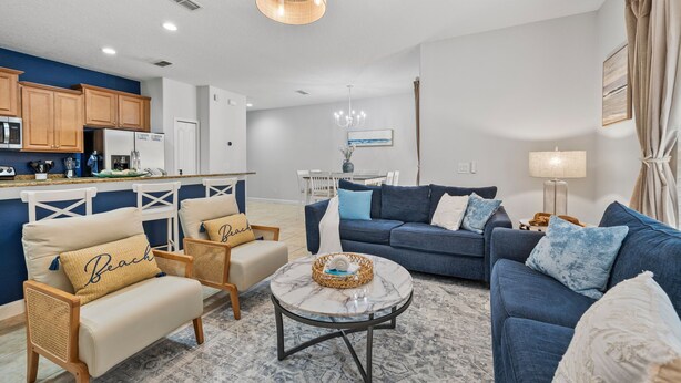 A living room with a blue sofa, two armchairs, a coffee table, and a kitchen area with wooden cabinets and a refrigerator. - NEW! Minutes to Disney World, Themed, Private Pool, Poolside Bar, Movie Theatre! (Kissimmee)