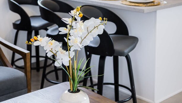 Una cocina moderna con una isla central, taburetes negros, un jarrón blanco con flores e iluminación colgante de mimbre.