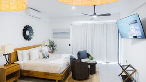 A modern hotel room with a bed, a TV, a ceiling fan, and a large window with curtains. - Vista Pedregal Comfortable Apartment in Cabo San Lucas Golden Zone (Cabo San Lucas)