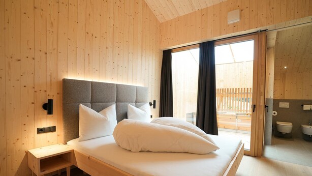 A bedroom with a wooden headboard, a bedside table, a bed with pillows, and a view of an outdoor area through a sliding door. - Vrbo Property (Prateria)