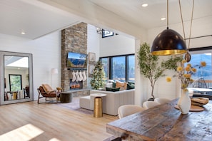 A modern living room with a stone fireplace, large windows, and a wooden dining table. - Nobby Duck Hollow (Gatlinburg)
