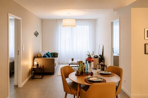 Un salon moderne avec un canapé, une table à manger dressée pour deux personnes et une grande fenêtre ornée de rideaux légers.