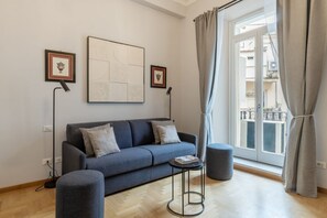 Ein modernes Wohnzimmer mit einer Couch, einem Hocker und einem kleinen Tisch. An der Wand befinden sich gerahmte Bilder, ein Fenster mit Vorhängen und ein Blick ins Freie.