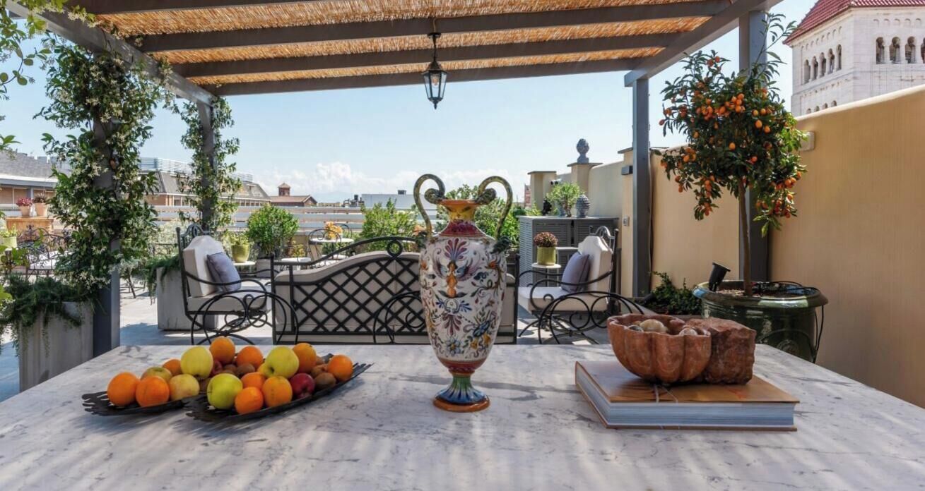 A table with a vase, fruits, and a book on a terrace with a view of buildings.