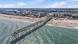 Zona costera con un muelle que se adentra en el agua, edificios frente a la playa y un paisaje urbano distante.