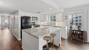 Una cocina moderna con armarios blancos, un refrigerador negro, una isla de mármol con taburetes y un mueble de madera con cristalería.
