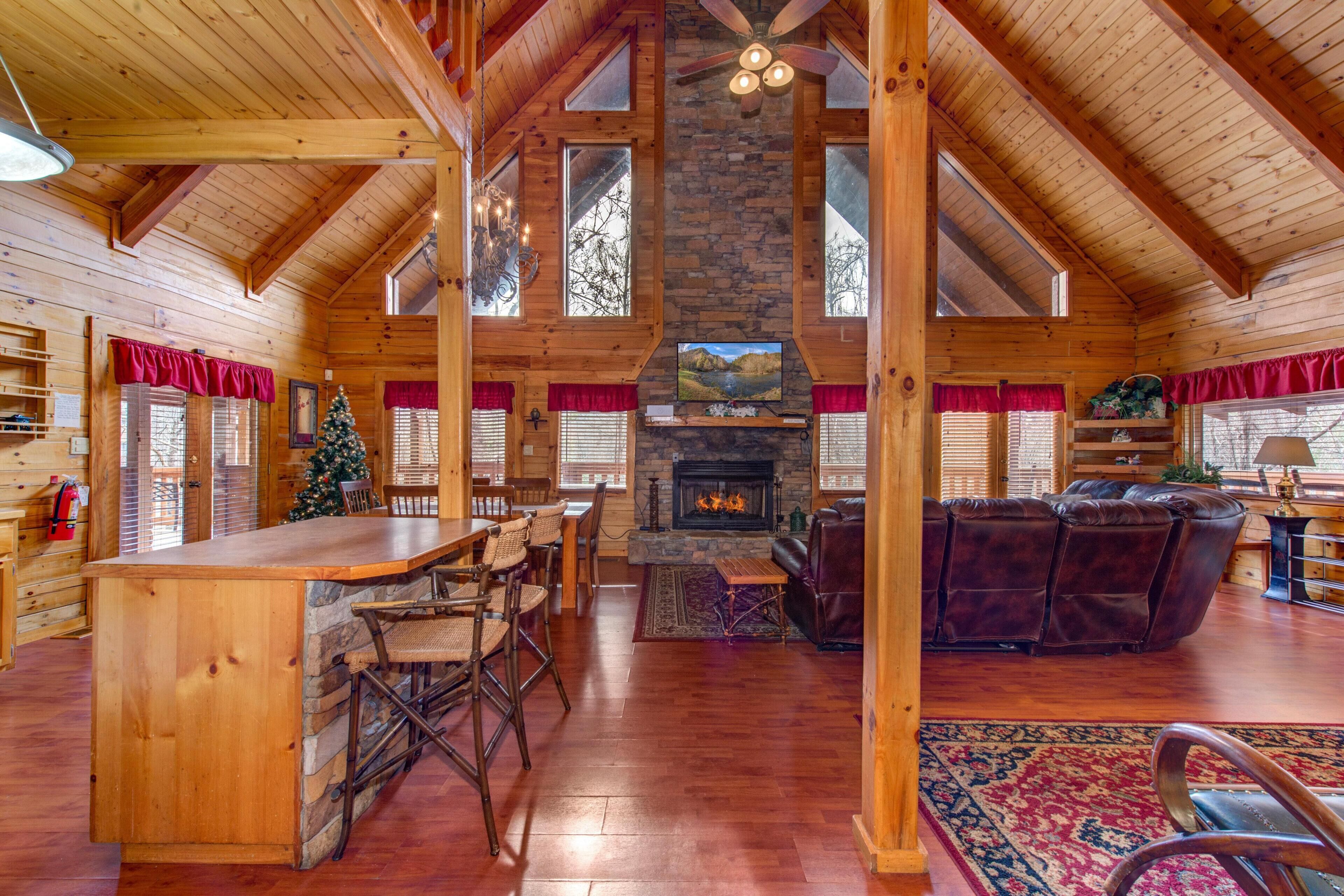 A spacious log cabin interior with a stone fireplace, wooden ceiling beams, a dining area, and a cozy seating area.