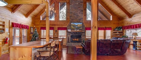 A spacious log cabin interior with a stone fireplace, wooden ceiling beams, a dining area, and a cozy seating area.