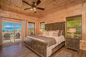 Quarto de cabana de madeira com uma cama, mesas de cabeceira, ventilador de teto e janelas amplas.
