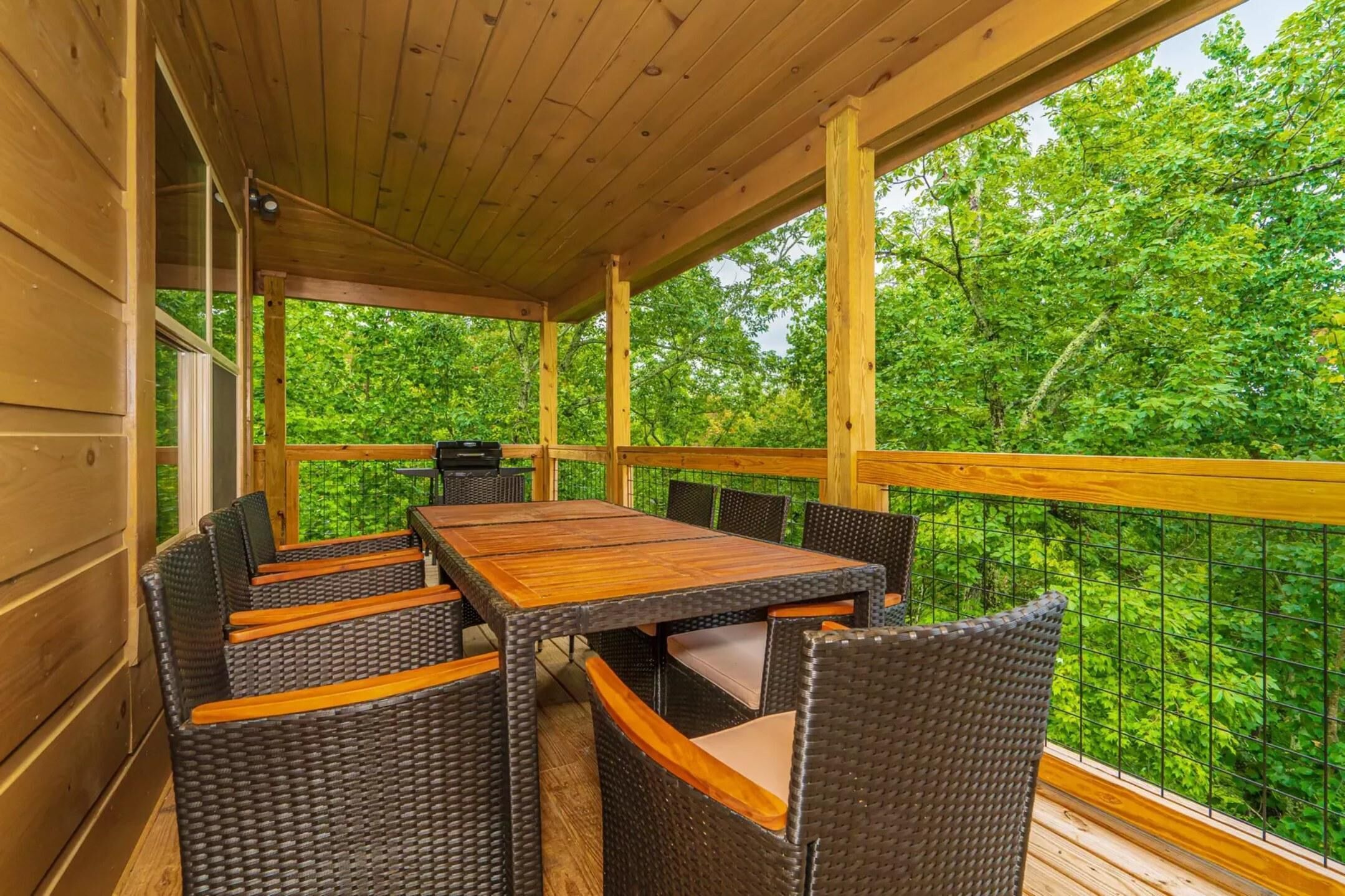 Un espace extérieur abrité, aménagé pour les repas, avec une table en bois et des chaises en osier, entouré d’une balustrade en bois offrant une vue sur une forêt dense et verdoyante.
