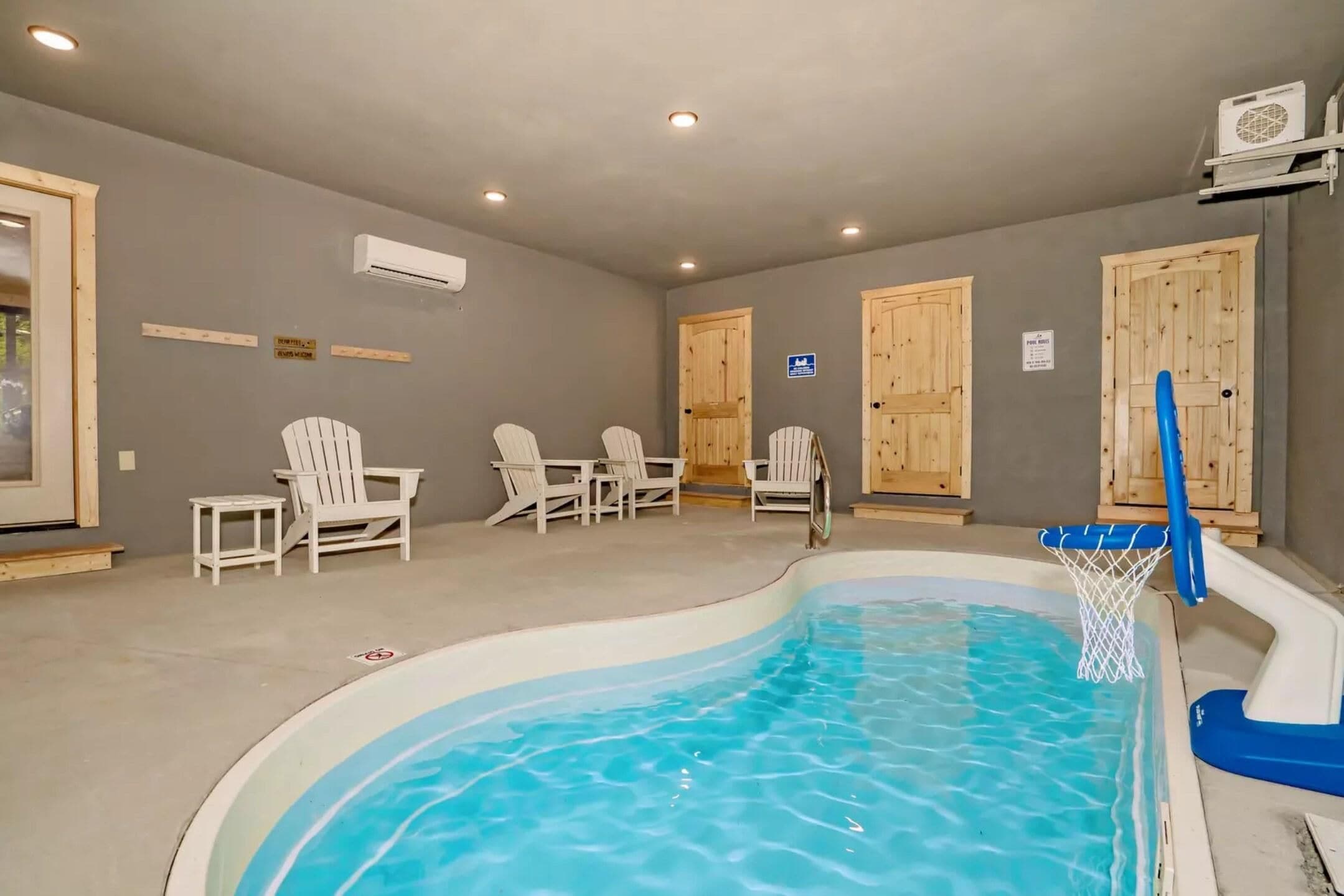 A swimming pool with a basketball hoop, white chairs, and wooden doors.