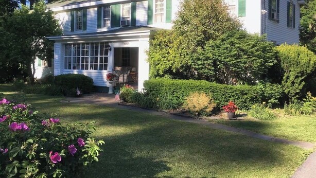 Una casa de dos pisos con un porche delantero, rodeada de jardines y árboles bien cuidados.