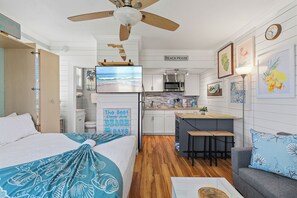 Ein kompaktes Hotelzimmer mit Küchenzeile, einem Bett mit Stranddecke, Deckenventilator und einem Schild, das anzeigt, dass das Zimmer 'Beach House' heißt.