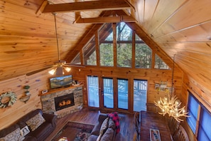 A log cabin interior with a stone fireplace, wooden ceiling beams, a large window, and a dining area. - A Grand Getaway (Gatlinburg)