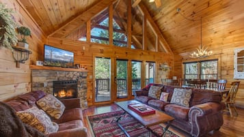 A log cabin interior with a fireplace, leather sofas, a dining area, and a large window.