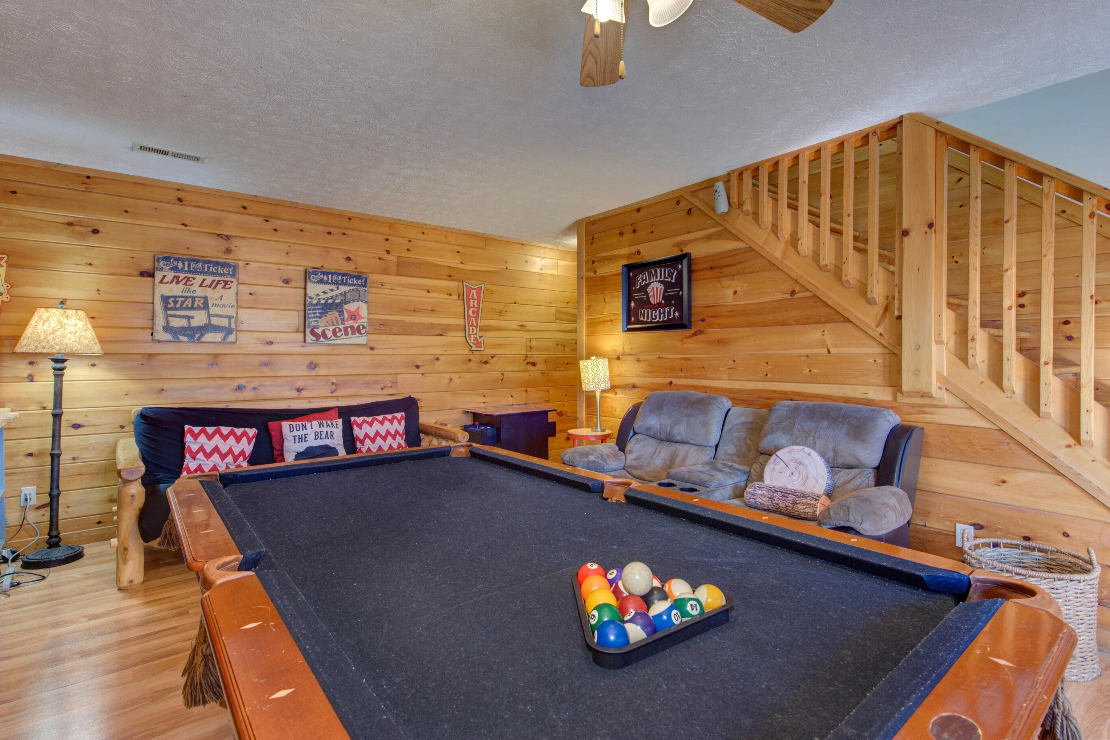 A pool table with balls, a sofa with cushions, a lamp, and a wooden staircase.