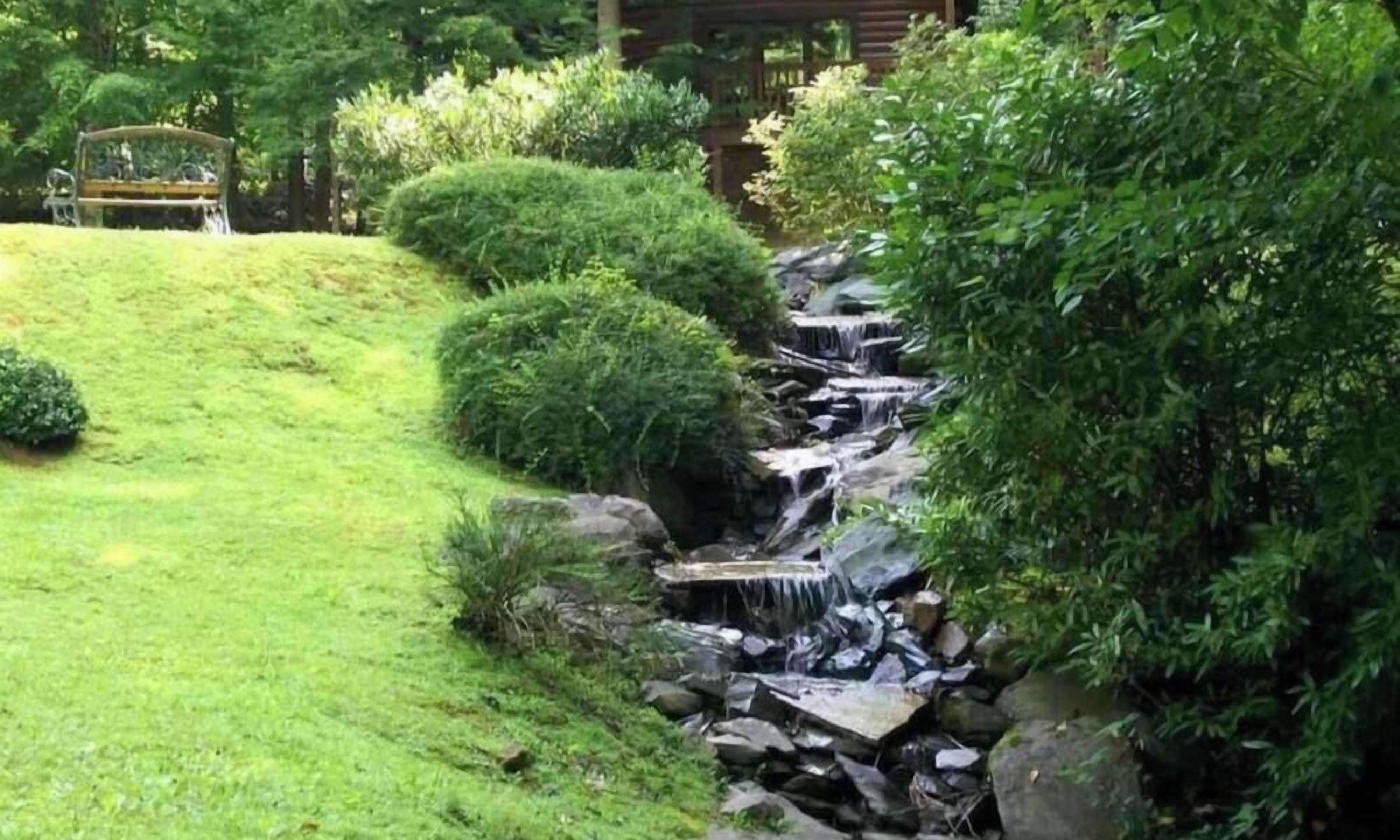 A garden with a small waterfall, neatly trimmed bushes, and a wooden bench.