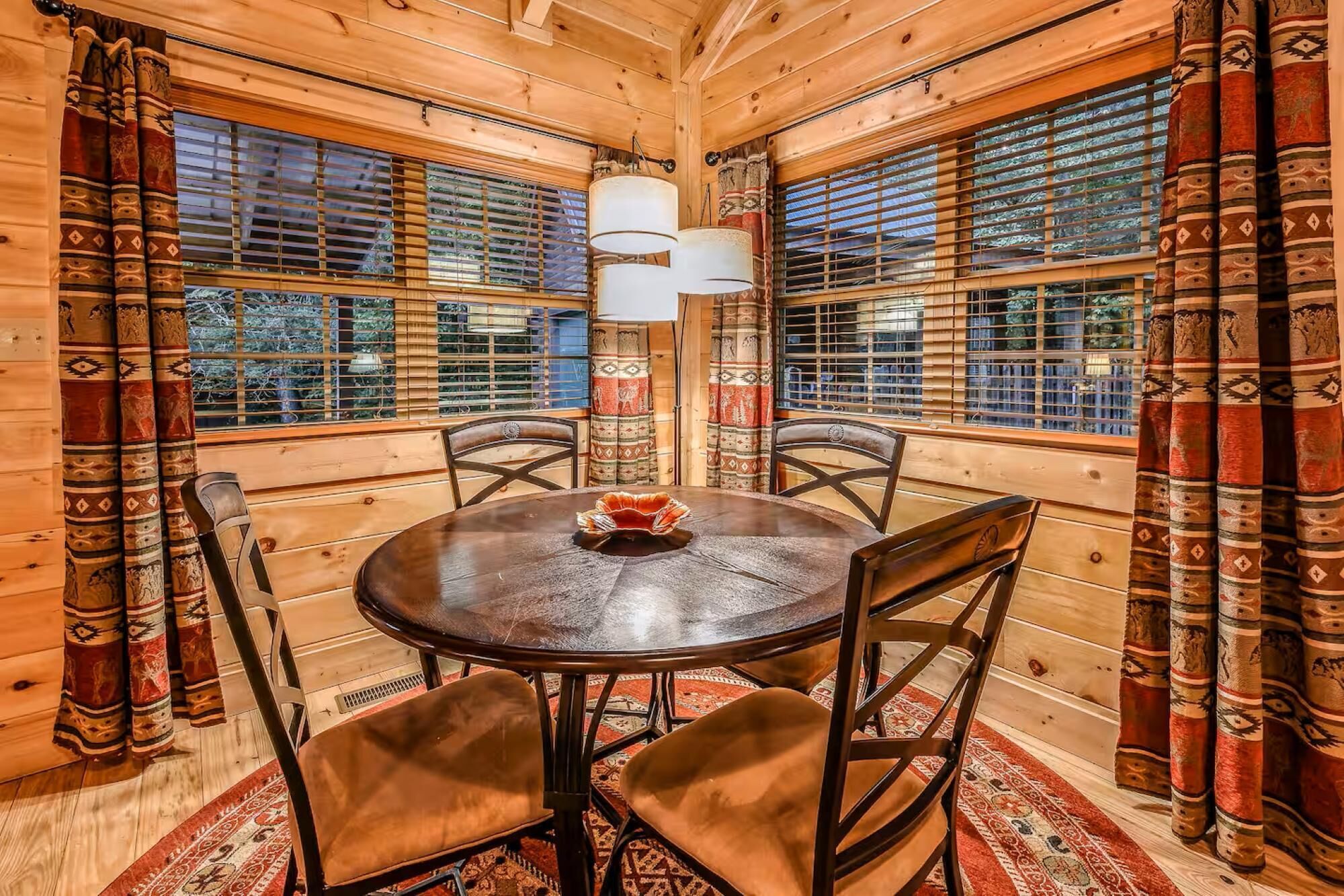 Un interior de madera con una mesa redonda, cuatro sillas, cortinas estampadas y una ventana con persianas.