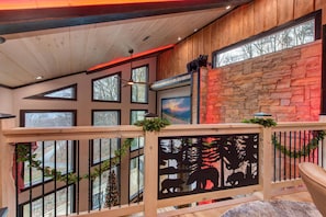 A wooden interior with a stone wall, multiple windows, and a balcony railing with a nature-themed cutout. - Waterfalls and Wildflowers (Gatlinburg)