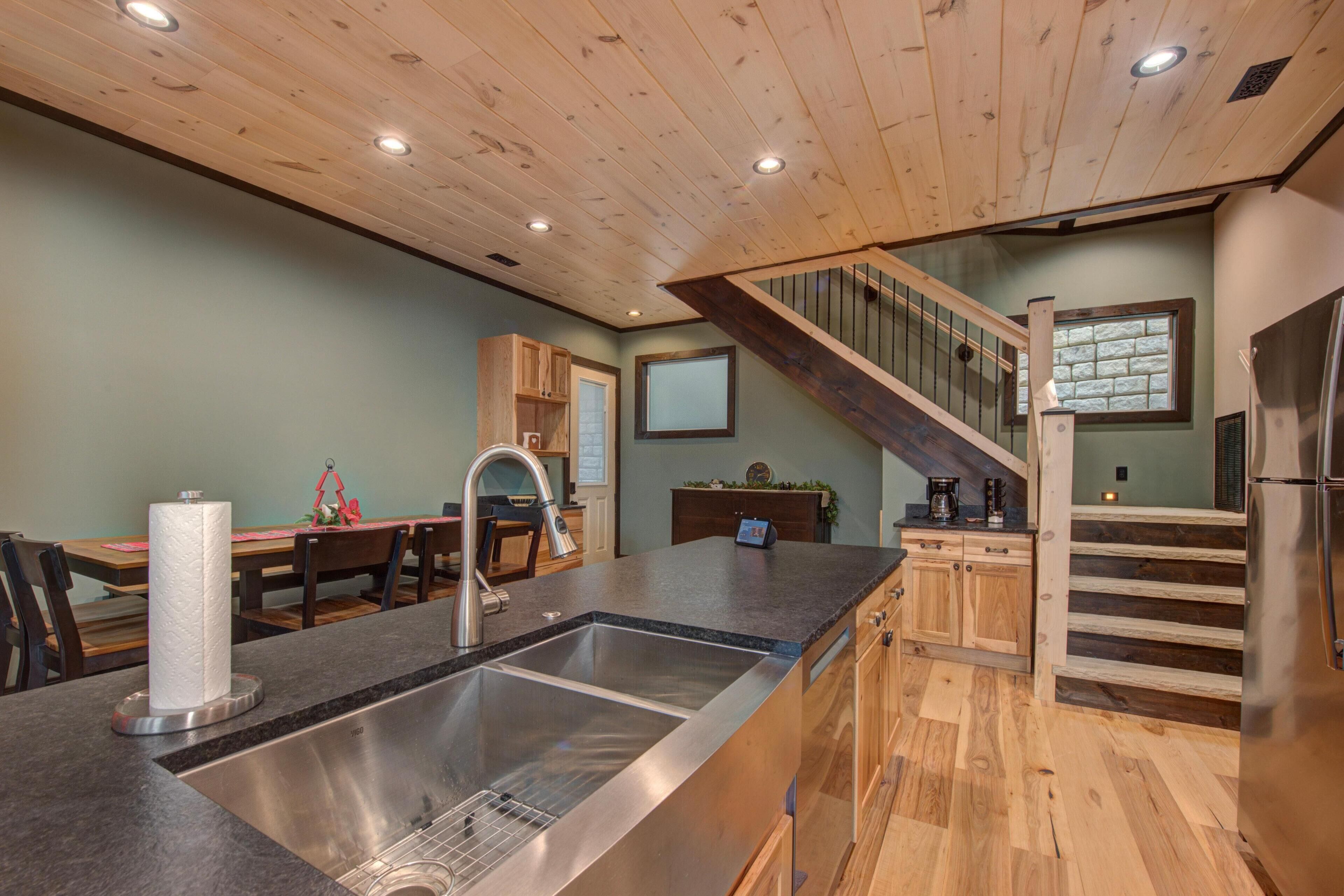 A kitchen with a stainless steel sink, wooden cabinets, a dining area with a table and chairs, and a staircase leading to an upper level.