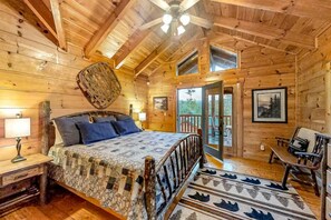A wooden cabin bedroom with a bed, a ceiling fan, a window, and a door leading to an outdoor area. - Bear Tracks (Sevierville)