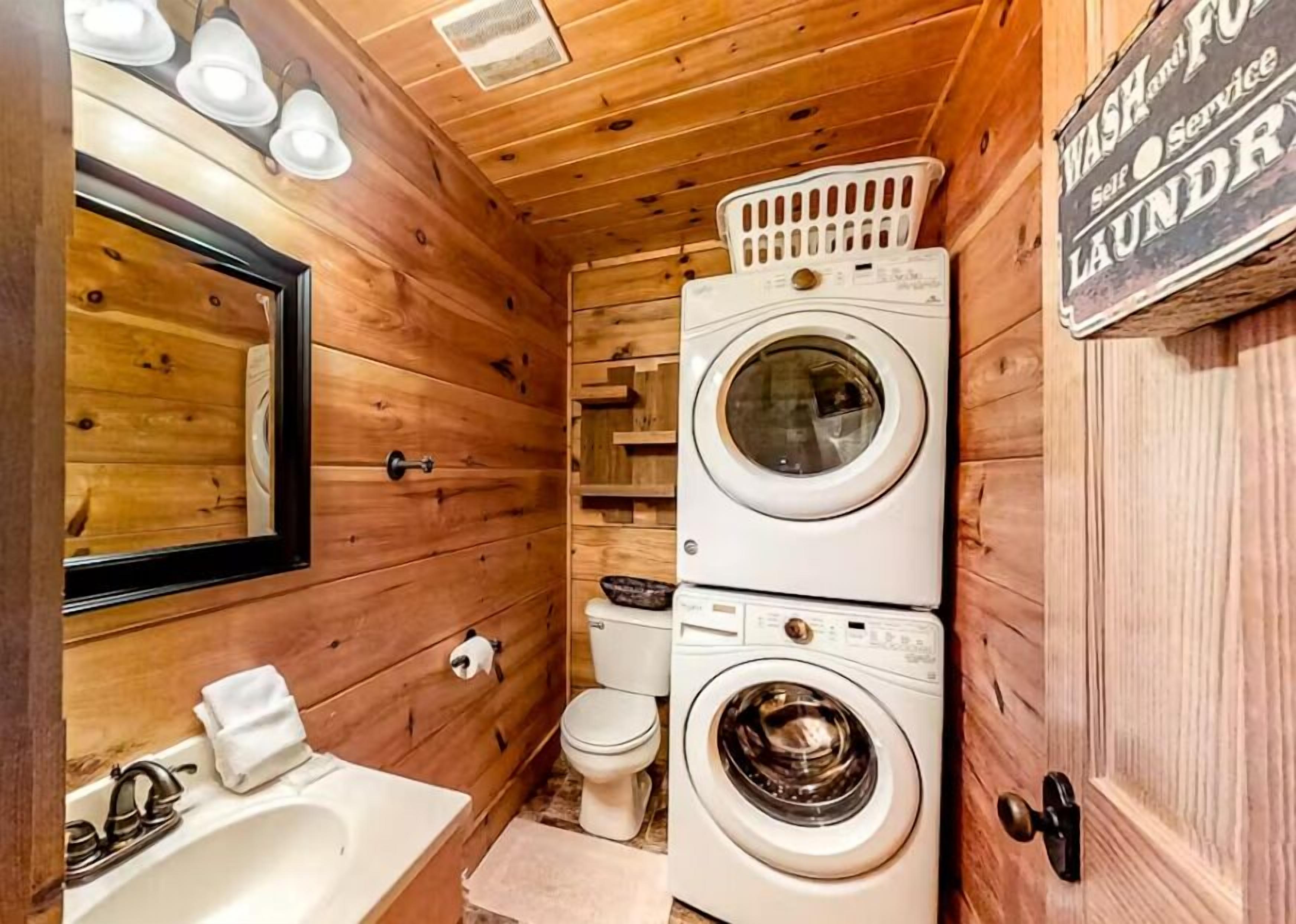 A compact, wooden-themed bathroom with a sink, mirror, two stacked washing machines, and a toilet.
