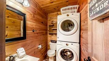 A compact, wooden-themed bathroom with a sink, mirror, two stacked washing machines, and a toilet.