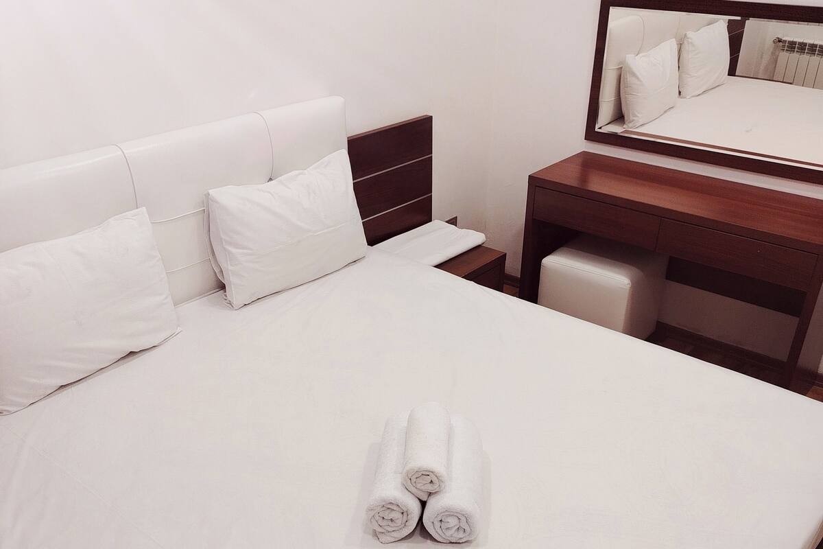 A neatly made bed with white linens and pillows, a wooden nightstand with a mirror, and a wooden floor.