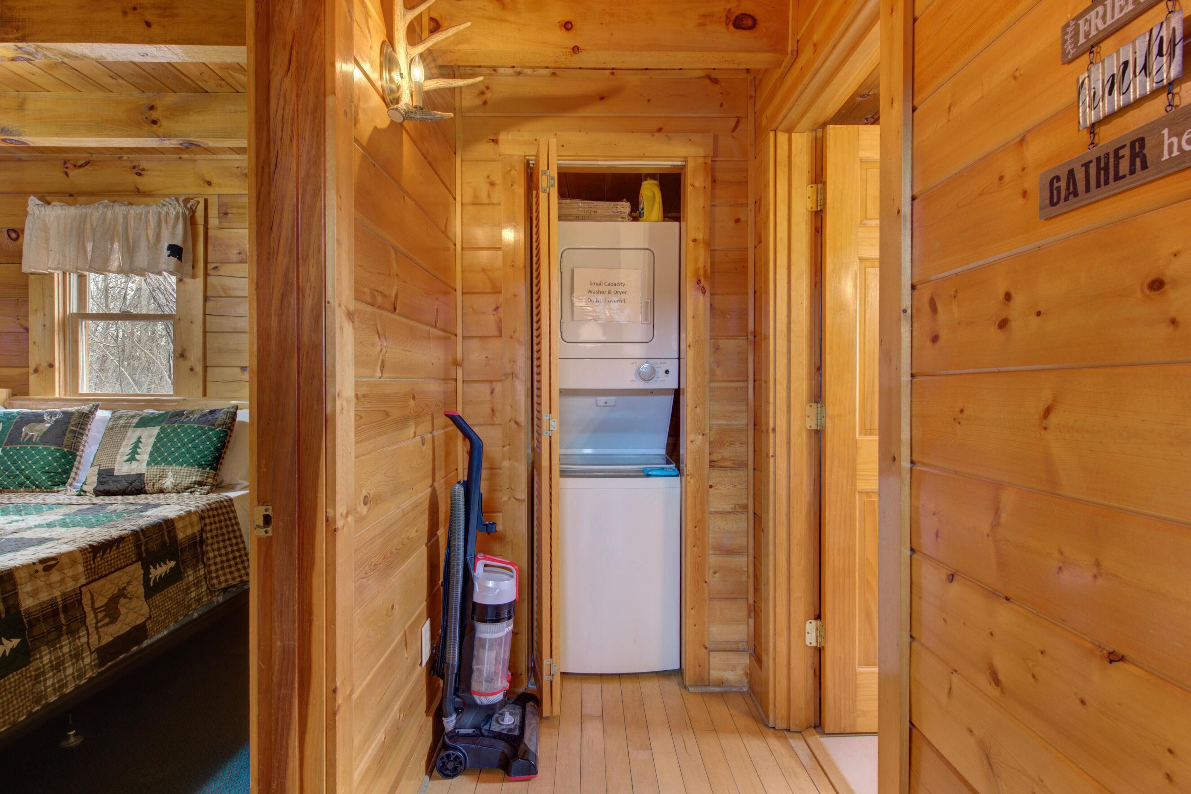 A compact room with a bed, a vacuum cleaner, a small refrigerator, and a wooden wall with a 'GATHER HERE' sign.