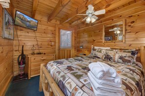 A wooden cabin bedroom with a bed, a ceiling fan, a television, and a window with blinds. - Bearcation (Pigeon Forge)
