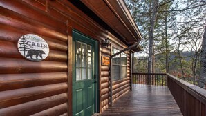 Une cabane en bois avec une porte verte, une pancarte indiquant « Bienvenue », et une vue sur les arbres.