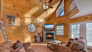 L’intérieur d’une cabane en rondins, avec une cheminée, des murs en bois, un ventilateur de plafond et une grande fenêtre.