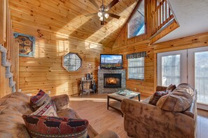 A log cabin interior with a fireplace, wooden walls, a ceiling fan, and a large window. - Lil Smoky Mountain Retreat (Sevierville)