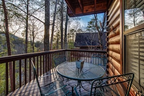 A wooden deck with a round metal table and chairs, surrounded by trees. - Lil Smoky Mountain Retreat (Sevierville)