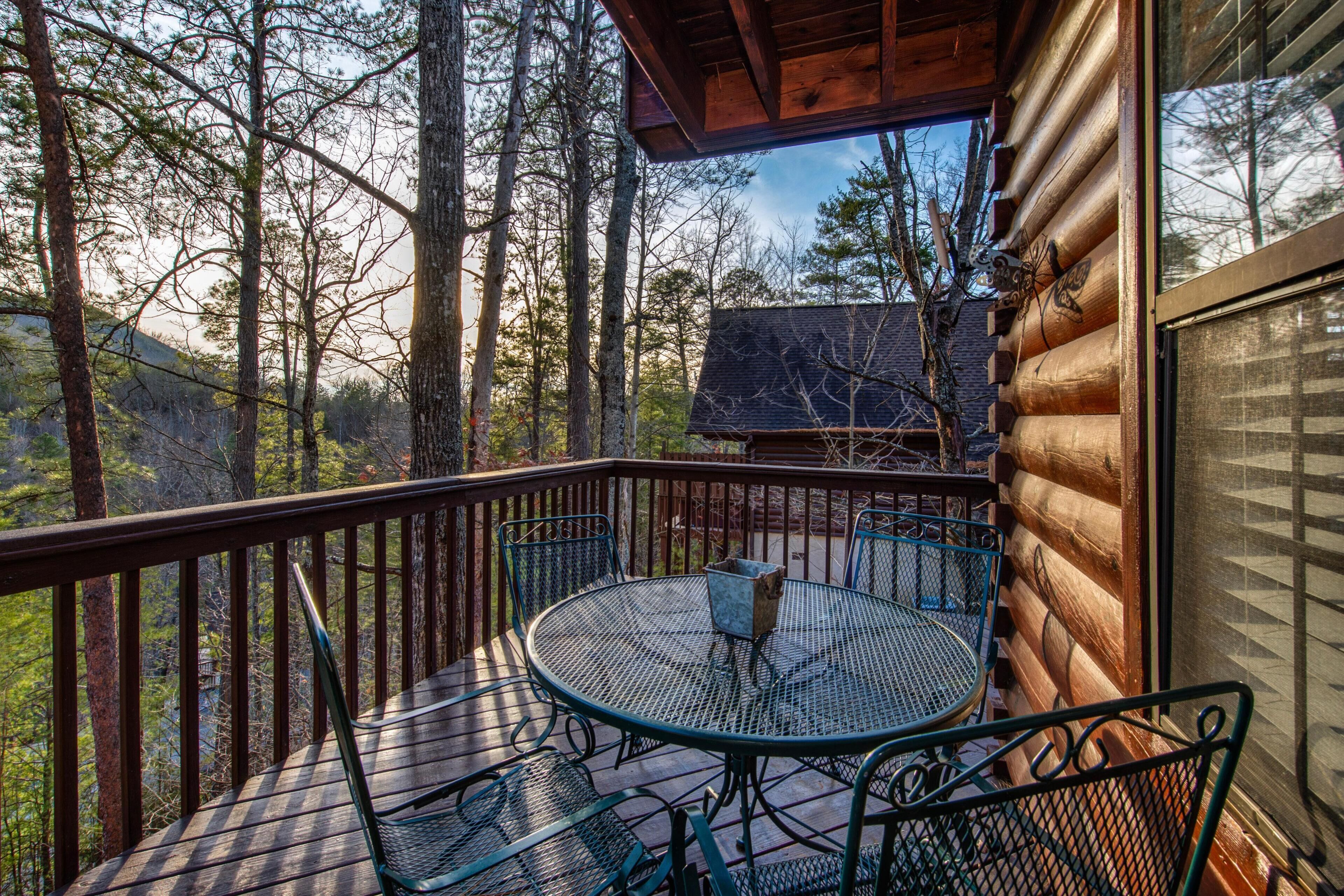 A wooden deck with a round metal table and chairs, surrounded by trees.