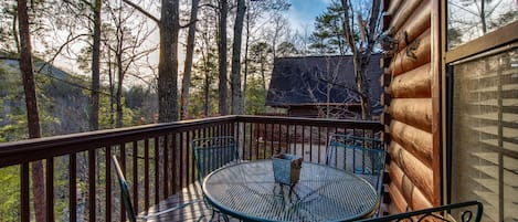 A wooden deck with a round metal table and chairs, surrounded by trees.