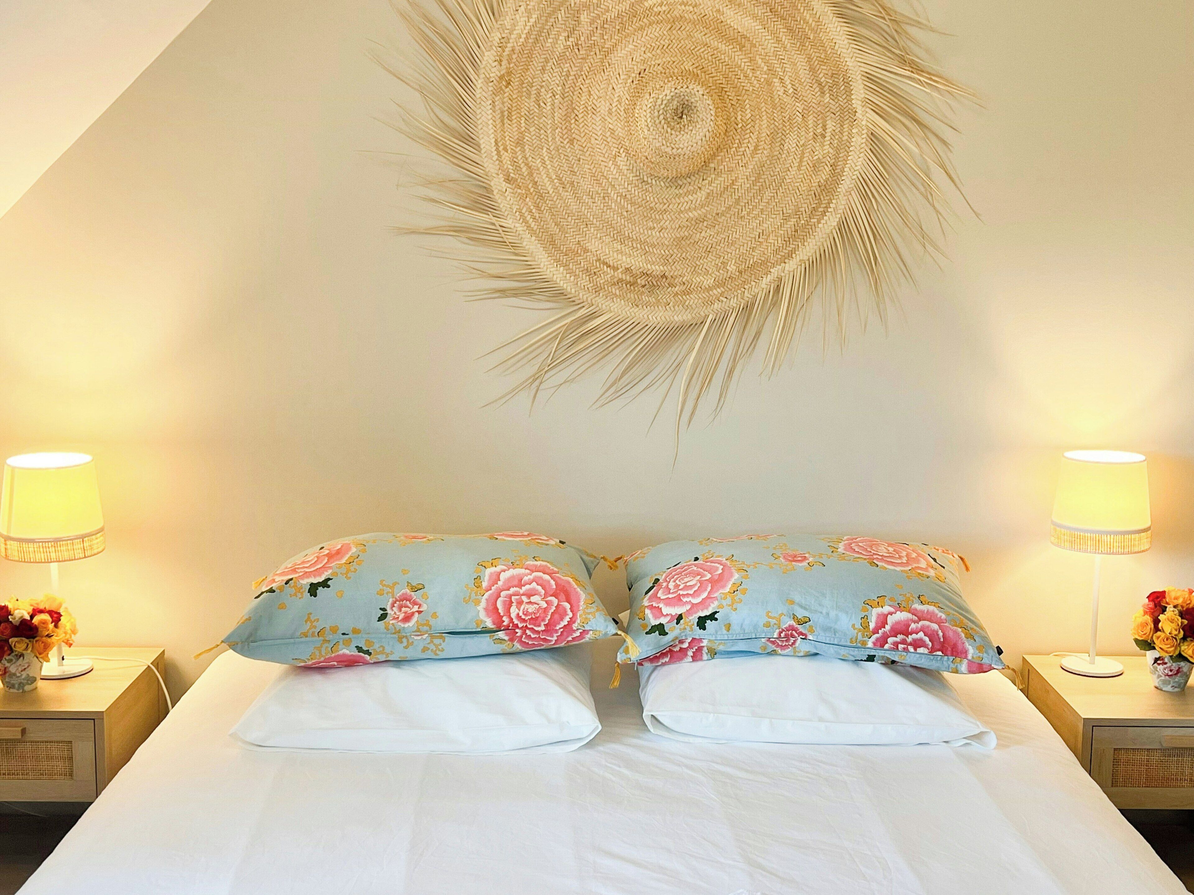 Un letto con due cuscini, due abat-jour, un pannello decorativo intrecciato e un piccolo vaso con fiori su un comodino.