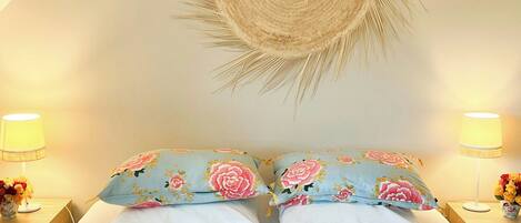 Un letto con due cuscini, due abat-jour, un pannello decorativo intrecciato e un piccolo vaso con fiori su un comodino.