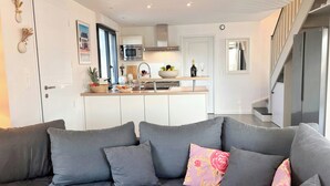 A modern living room with a grey sofa, a kitchen in the background, and a staircase leading to an upper level. - Beachside villa with jacuzzi in Brignogan (Brignogan Plages)