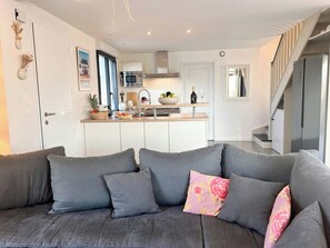Ein modernes Wohnzimmer mit einem grauen Sofa, einer Küche im Hintergrund und einer Treppe, die in einen oberen Bereich führt.
