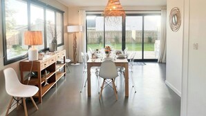 A dining area with a table set for four, a modern lamp, and a view of the backyard through large windows. - Beachside villa with jacuzzi in Brignogan (Brignogan Plages)