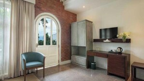 Una habitación con pared de ladrillo, puerta blanca, silla azul, escritorio, televisor y una pequeña mesa con un florero.