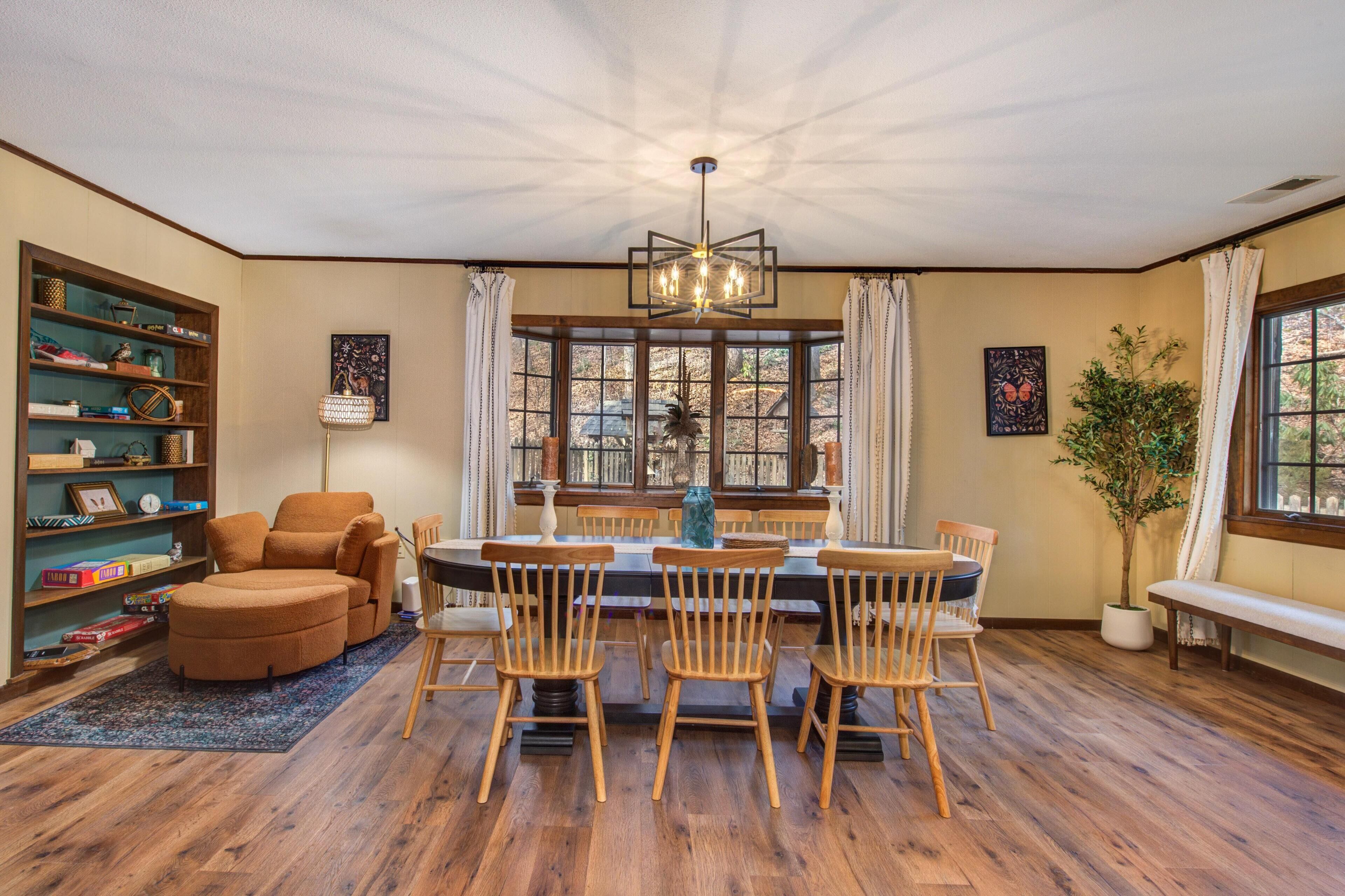 A dining area with a round table, wooden chairs, a bookshelf, and a large window with curtains.