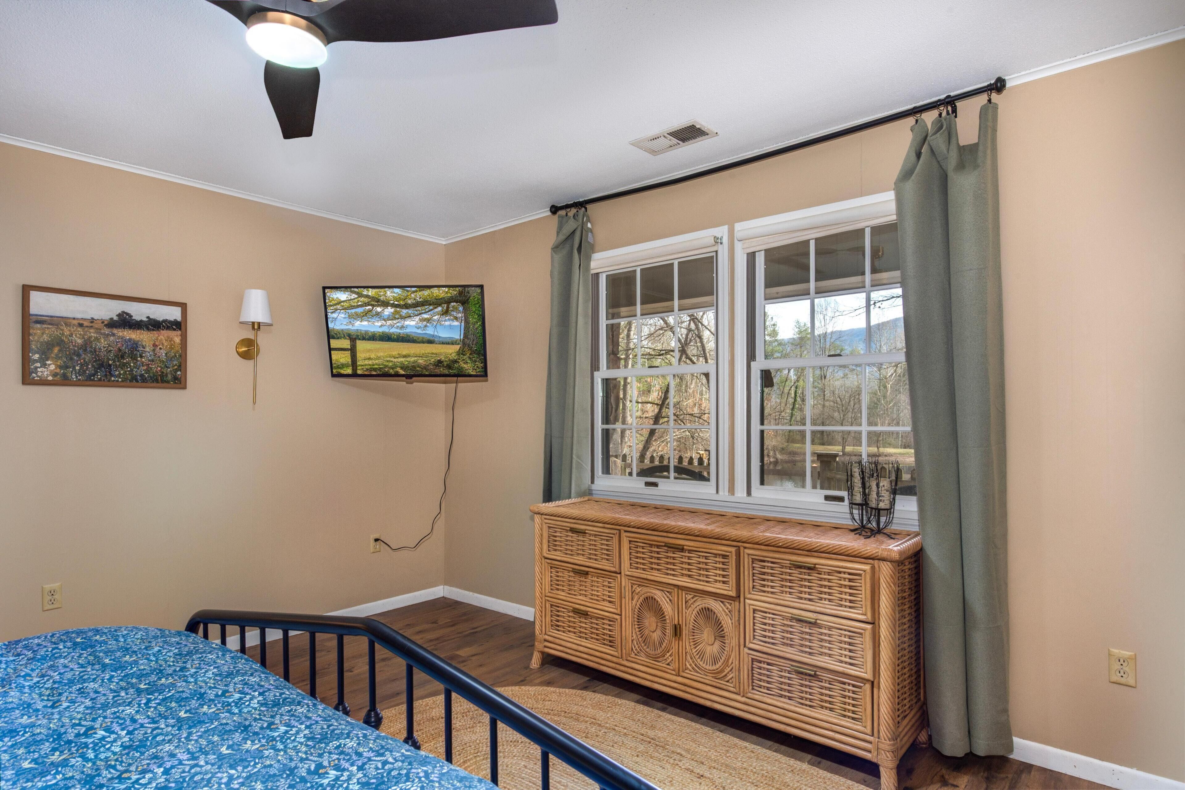 Une chambre à coucher comprenant un lit, une commode, une fenêtre avec des rideaux, un ventilateur de plafond et des œuvres d’art accrochées au mur.