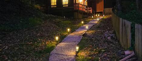A pathway leading to a wooden building with illuminated windows and a wooden fence.