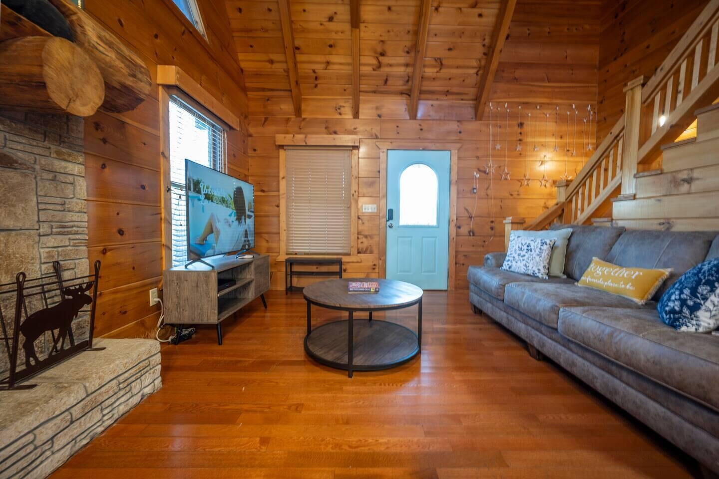 A log cabin interior with a wooden floor, a sofa, a round coffee table, a television, and a stone fireplace.
